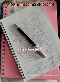 Two spiral-bound notebooks—one pink and one white—rest on a surface. The pink notebook is labeled “ColourMine Pink Note Pad.” The white notebook is open, displaying handwritten cursive notes with a black and silver pen laid diagonally across the page. Copyright marks “© Susan Lane 2025” and “© Susan Lane  ColourMine.com” are visible.
