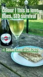 A green colour bottle of Martini sparkling wine, a filled glass, and a plate with buttered Ryvita's on a wooden outdoor table, set against a green lawn and fence. Overlaid text reads: “Colour  This is love, hues, SEO survival © Susan Lane SLCM 2025.