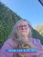 A woman with long light hair and glasses sits outdoors in front of a tall green hedge, wearing a pink top, with the text ‘Susan Lane SLCM 2025’ in the corner.